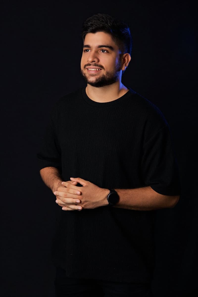 Juan Camilo Rojas smiling, standing with his hands together against a black background with blue side lighting